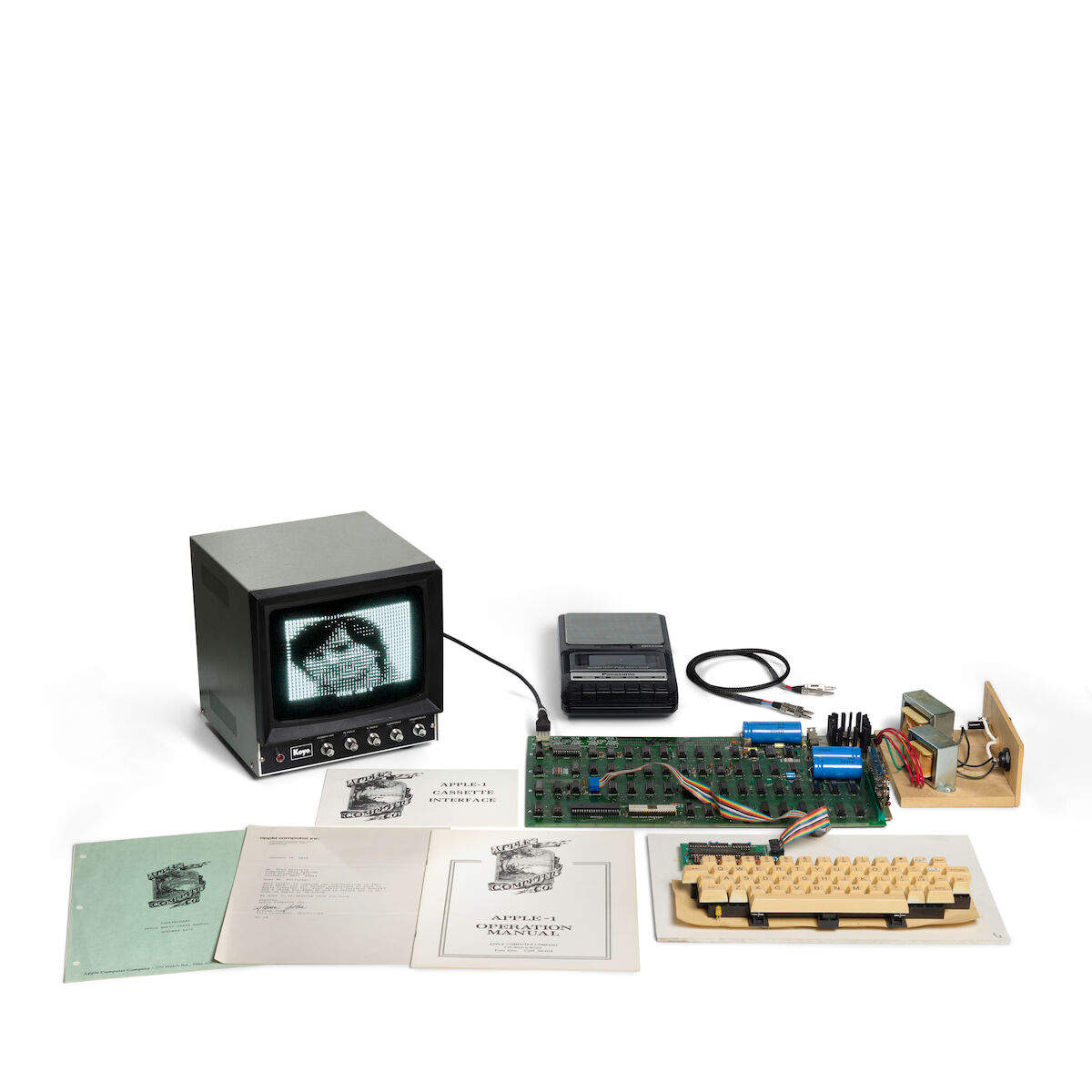 La carte mère de l'Apple-1 sur fond blanc, avec un petit écran, un clavier, l'interface cassette et des manuels.