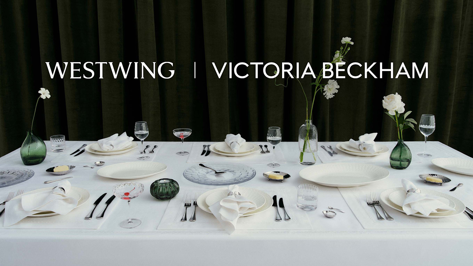 Une longue table dressée de blanc devant un rideau vert sombre, avec fleurs, verres, assiettes et couverts, sous la mention « Westwing | Victoria Beckham ».