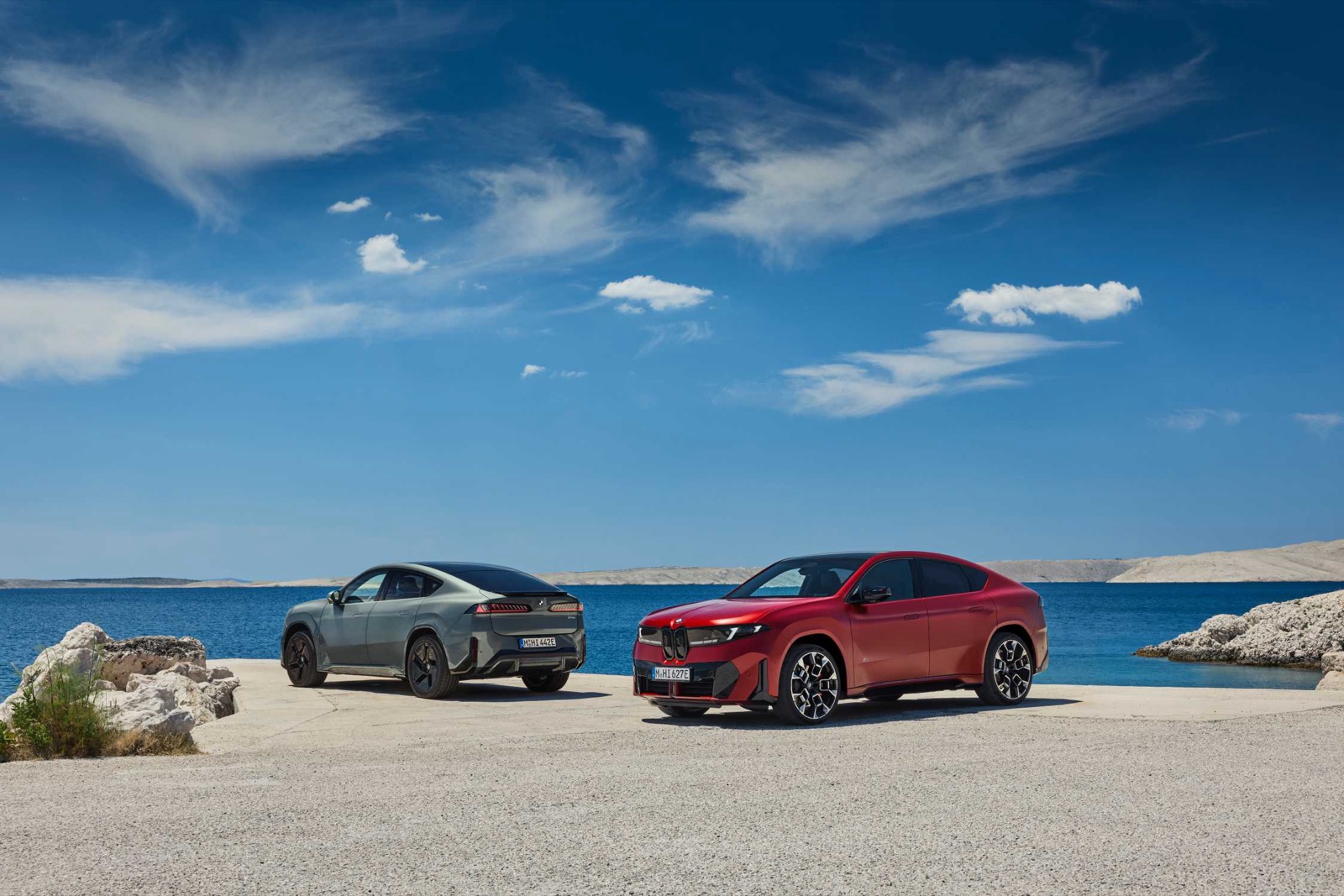 Deux BMW iX4, l'une rouge de trois quarts avant, l'autre grise de trois quarts arrière, garées au bord d'une mer sous un ciel bleu.