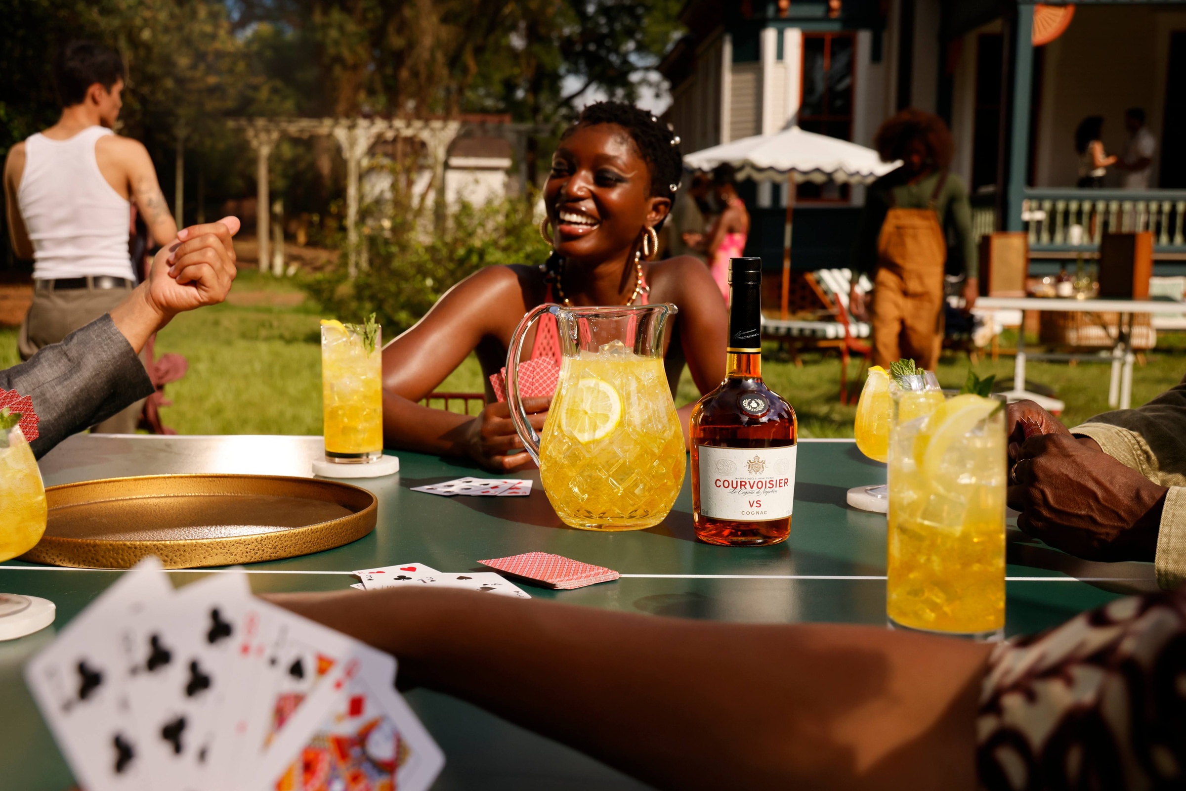 Table de jardin avec des cartes à jouer, des verres de cocktail et une bouteille de Courvoisier VS, devant une femme qui rit