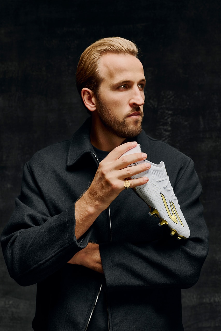 Harry Kane de profil sur fond sombre, tenant un crampon blanc et or et portant une chevalière en or.