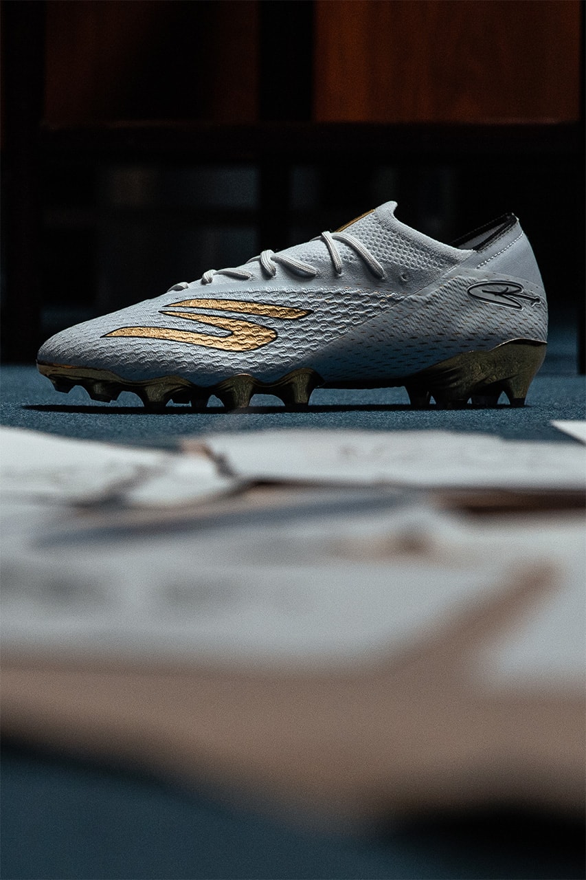 Crampon blanc à logo S doré et semelle dorée, photographié de profil sur une moquette bleue.
