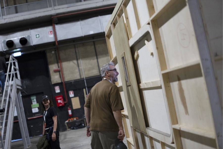 Un homme aux cheveux gris regarde un décor en bois sur un plateau de tournage, une femme derrière lui