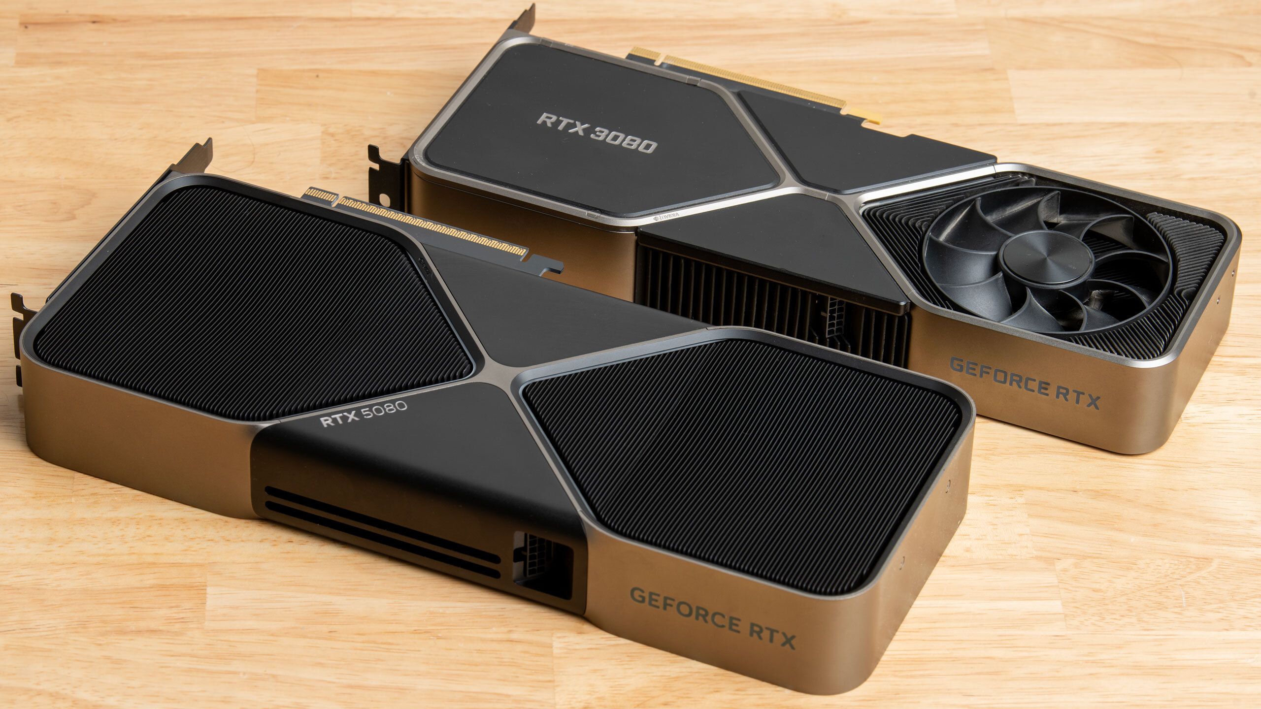 Une RTX 5080 Founders Edition posée à côté d'une RTX 3080 sur un plan de bois clair.