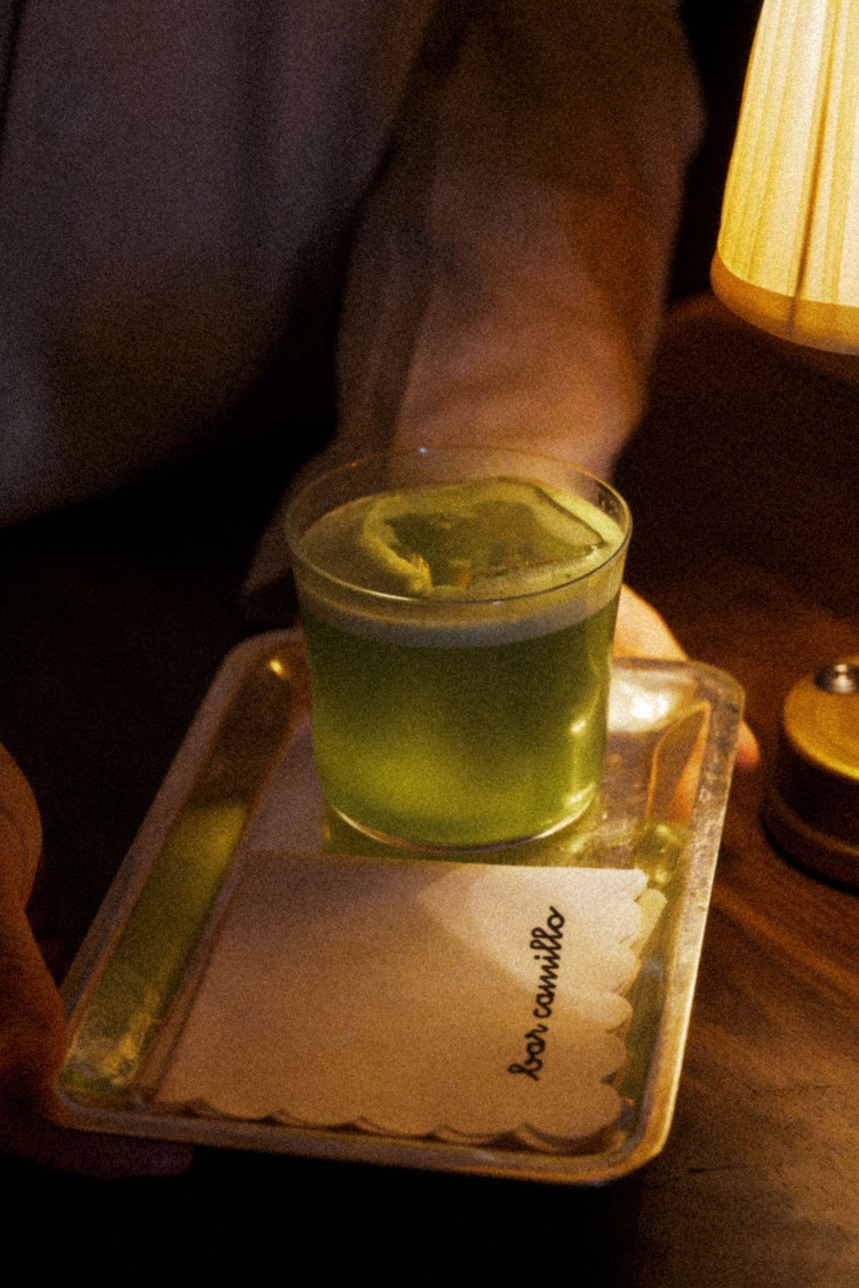 Un serveur tient un plateau argenté portant un cocktail vert sur glace et une serviette blanche festonnée marquée bar camillo.