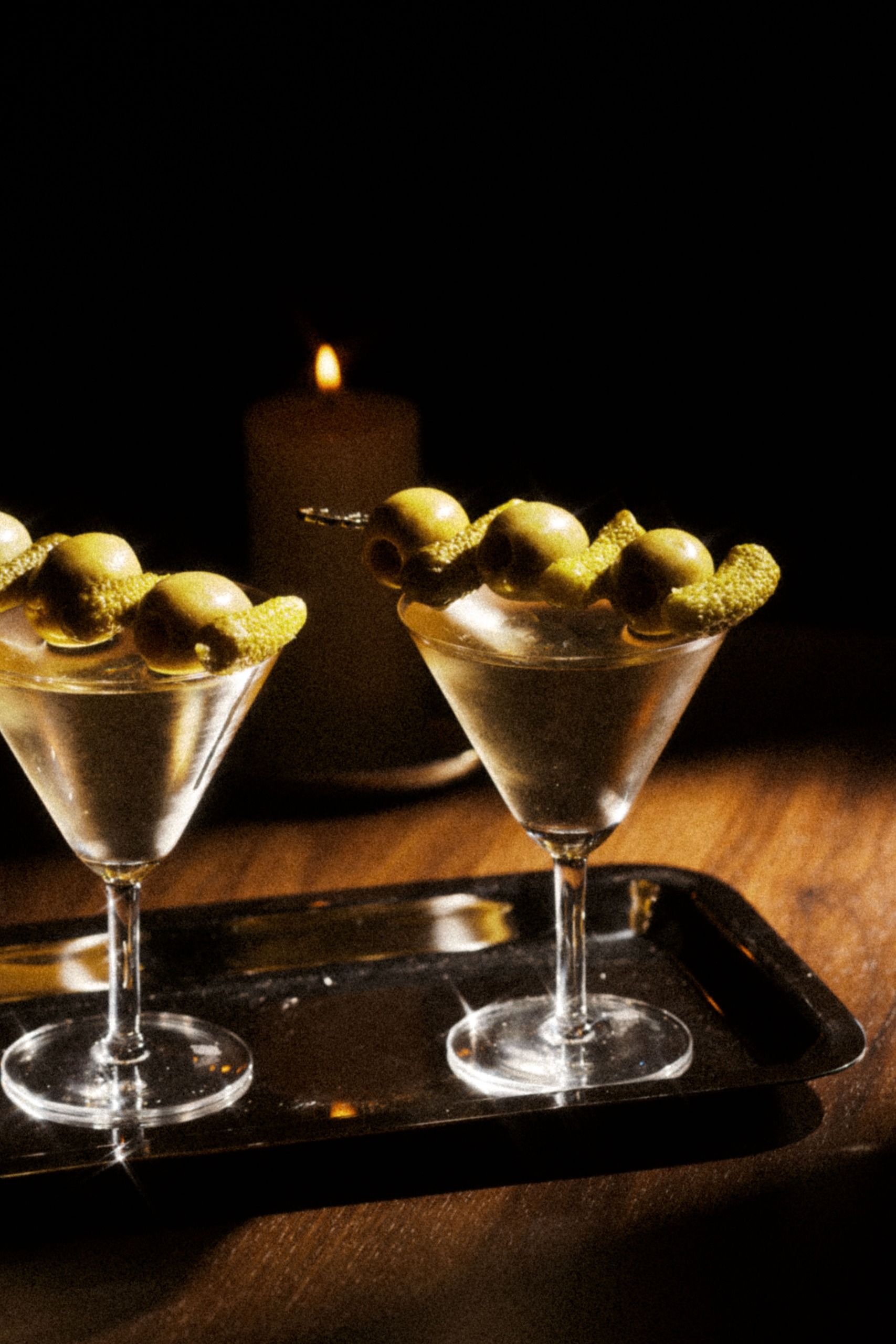 Deux verres à martini garnis d'olives et de zestes sur un plateau métallique, devant une bougie allumée.