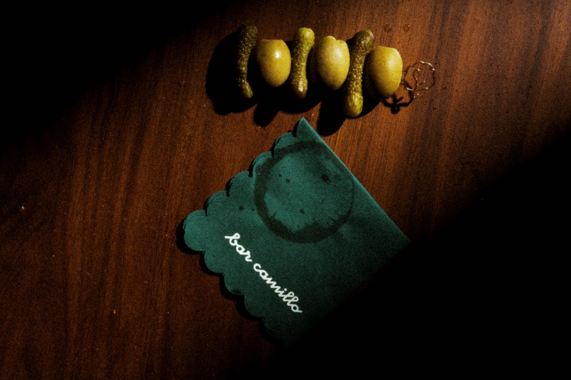 Serviette vert foncé à bord festonné portant l'inscription bar camillo, posée sur une table en bois sous trois olives et cornichons piqués.